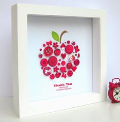 an apple made out of buttons in a white shadow box with a red alarm clock