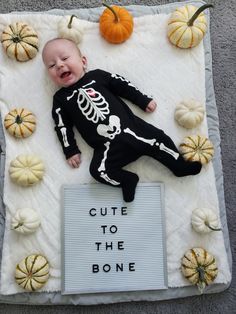 a baby in a skeleton costume laying on a sign with pumpkins around it that says cute to the bone