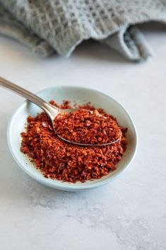 a white bowl filled with red spices next to a gray dish towel and silver spoon