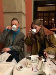 two people sitting at a table drinking from mugs in front of each other,