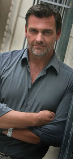 a man standing with his arms crossed in front of a metal fence and building behind him