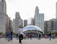 a woman standing in front of a large metal object with people walking around it and buildings behind her