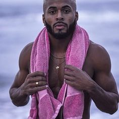 a shirtless man with a pink towel around his neck standing on the beach looking at the camera