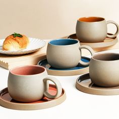 there are four cups and two plates on the table, one has a croissant in it