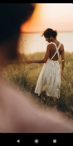 a woman in a white dress walking through tall grass with the sun setting behind her