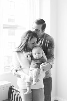 a man holding a baby while standing next to a woman in front of a window