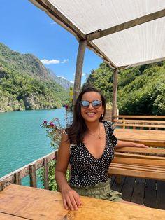 a woman sitting at a picnic table with mountains in the background and water behind her