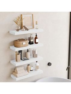 three white shelves with items on them against a wall in a bathroom, next to a bathtub