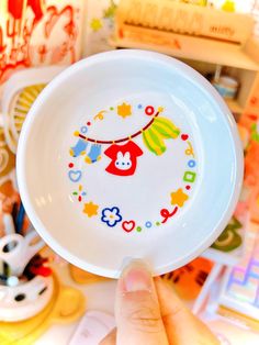 a hand holding a white bowl with colorful designs on the bottom and sides, in front of other children's toys