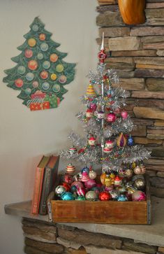 a small christmas tree with ornaments in a wooden crate next to a stone fireplace mantel