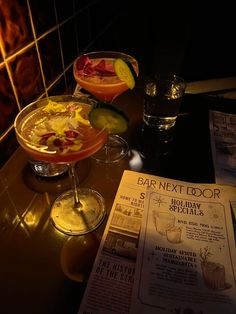 two cocktails sitting on top of a bar next to a menu and drinks glasses