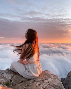 a woman sitting on top of a mountain with her hair blowing in the wind and clouds below