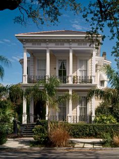 a large white house with columns and balconies on the second story, surrounded by palm trees