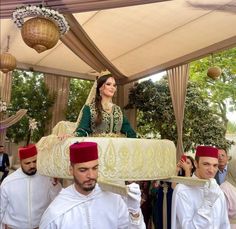 a man and woman in traditional garb carrying a large cake on their head while others look on