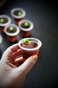 a hand holding a small cup filled with liquid and sprinkled with green olives