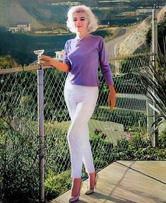 a woman in white pants and purple shirt standing on a wooden platform holding a cup