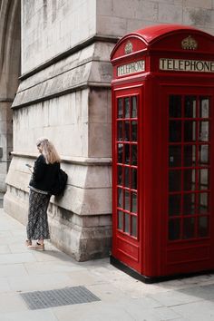 a woman leaning against a wall next to a red phone booth on the side of a building