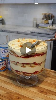 a person holding a spoon over a layered cake on a counter top with cans of milk in the background