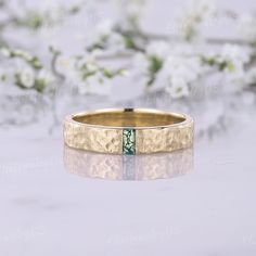 a gold wedding band with a green diamond in the center and flowers behind it on a reflective surface