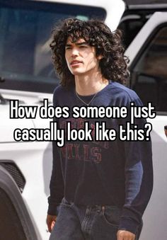 a man with curly hair wearing a black shirt and jeans standing in front of a white truck