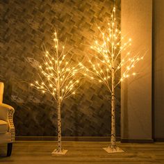 three lighted trees in the corner of a room with a chair and wall behind them