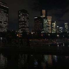 the city skyline is lit up at night with lights reflecting in the water and people walking around