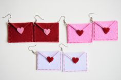 three small envelopes with hearts are hanging from hooks on a white wall, one is red and the other has pink felt