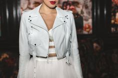 a woman wearing a white jacket and skirt with red lipstick on her lips is standing in front of a wall