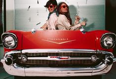 two women sitting on the hood of a red car with their arms around each other