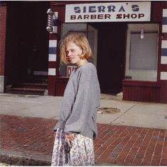 a woman is standing on the sidewalk in front of a barber shop with her hair pulled back