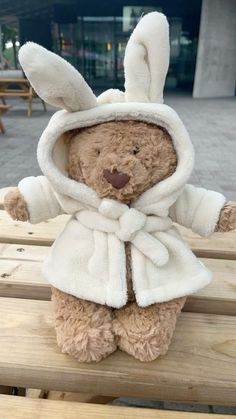 a teddy bear wearing a white coat and hoodie sitting on a wooden bench outside