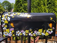 a black mailbox with yellow and white flowers painted on it's side next to a fence