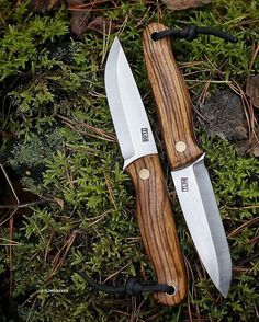 two knives sitting next to each other on top of green moss covered ground with trees in the background