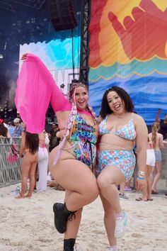 Two women stand side by side on sandy ground, posing in vibrant festival outfits. One wears a neon pink cape and a rainbow-patterned swimsuit, and the other wears a pastel floral bikini set. Behind them is a crowd and a large, colorful hand-shaped mural.