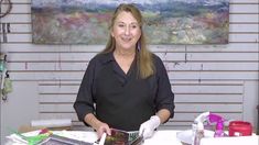 a woman standing in front of a table covered with art supplies and papers, smiling at the camera