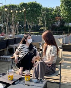 two women sitting on chairs wearing face masks and drinking beer at an outdoor bar with trees in the background