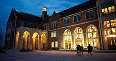 two people standing in front of a large building at night with lights on the windows