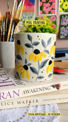 a coffee cup with yellow flowers painted on it sitting next to books and paintbrushes