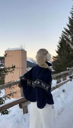 a woman standing in the snow looking out at mountains and trees, with her back to the camera