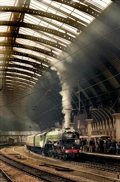 a train is coming down the tracks as people look on from inside an indoor station