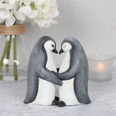 two penguins holding each other in front of a vase with flowers on the table next to it