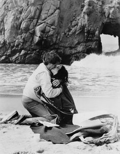 a man and woman hug on the beach near an arch in the rock formation behind them