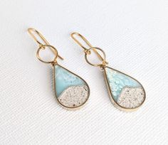 These lightweight teardrop dangle earrings, made from hand-colored eco-resin, feature a mix of crushed stones, silver specks, and real sand. The body and wiring are created with jewelers brass; for an additional cost, 14 K Gold Filled Ear Wiring is also available. The earrings measure 1.25" high and hang approximately 1.75" from the ear. For best results, each piece is handmade and crafted individually; slight variations of color and sand shapes should be expected. To maintain luster, please rem Coastal Jewelry, Wood Resin Jewelry, Beach Earrings, Resin Ideas, Eco Resin, Earrings Summer, Beach Necklaces