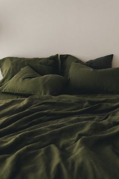 an unmade bed with dark green sheets and pillows in a white walled bedroom area