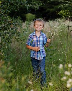 a young boy standing in tall grass holding onto a plant with one hand and smiling at the camera