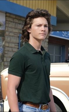 a young man standing in front of a car wearing a green shirt and blue jeans