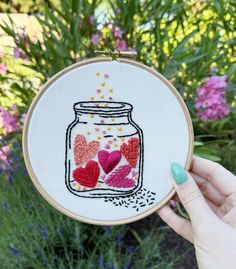 a hand holding up a embroidery project with hearts in a jar on the front and pink flowers behind it