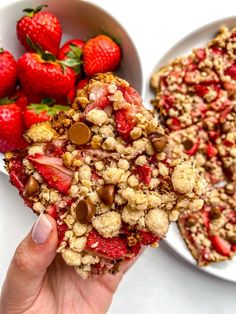 someone holding up a piece of strawberry crumbler cake next to two bowls of strawberries