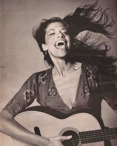 a woman with her hair blowing in the wind while holding an acoustic guitar and laughing