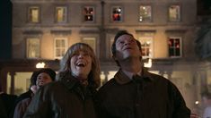 two people standing next to each other in front of a building at night with lights on